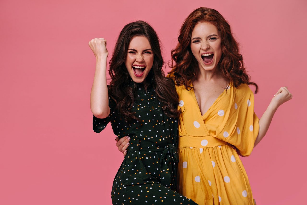 optimistic-women-in-bright-dresses-happily-shout-on-pink-background-joyful-girlfriends-with-curly